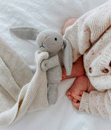 A baby is laying on a bed with a Bengali Collections Bunny Snuggly - Natural Marle.