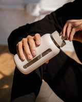 A person tracking their daily water needs with a Day Bottle with Hydration Tracker - Various Options by Bink on a wooden floor.