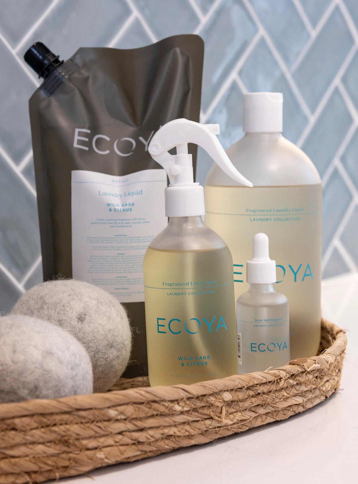 Ecoya refill pouch, linen spray, dropper and laundry liquid in a woven tray against blue tiles