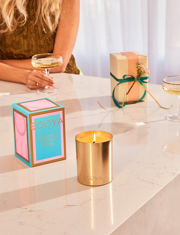 A woman sits at a table next to a Gift Box and an Ecoya Holiday: Blossom & Spiced Vanilla Goldie Candle.
