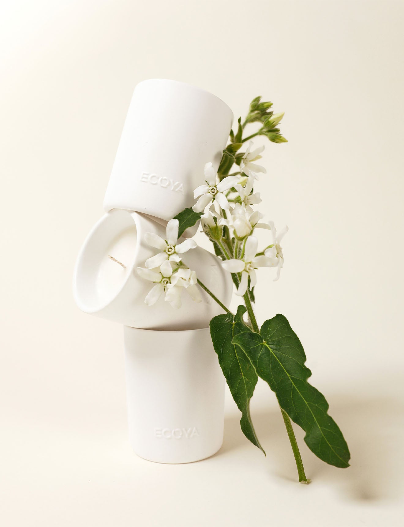Minimalist arrangement of three white ECOYA candles stacked in a sculptural composition, accented with fresh white flowers and green leaves, set against a soft cream background