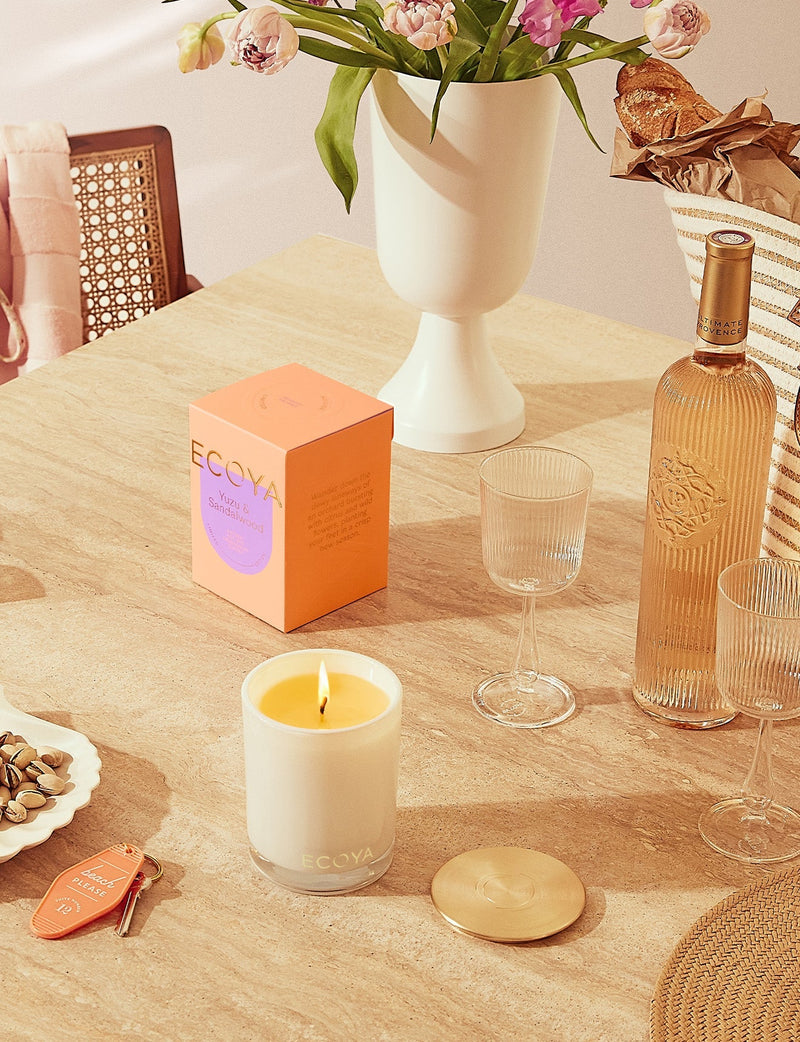 A Scandinavian-inspired Yuzu & Sandalwood Madison Candle by Ecoya, a perfect home design gift, placed on a table alongside a vase of flowers.