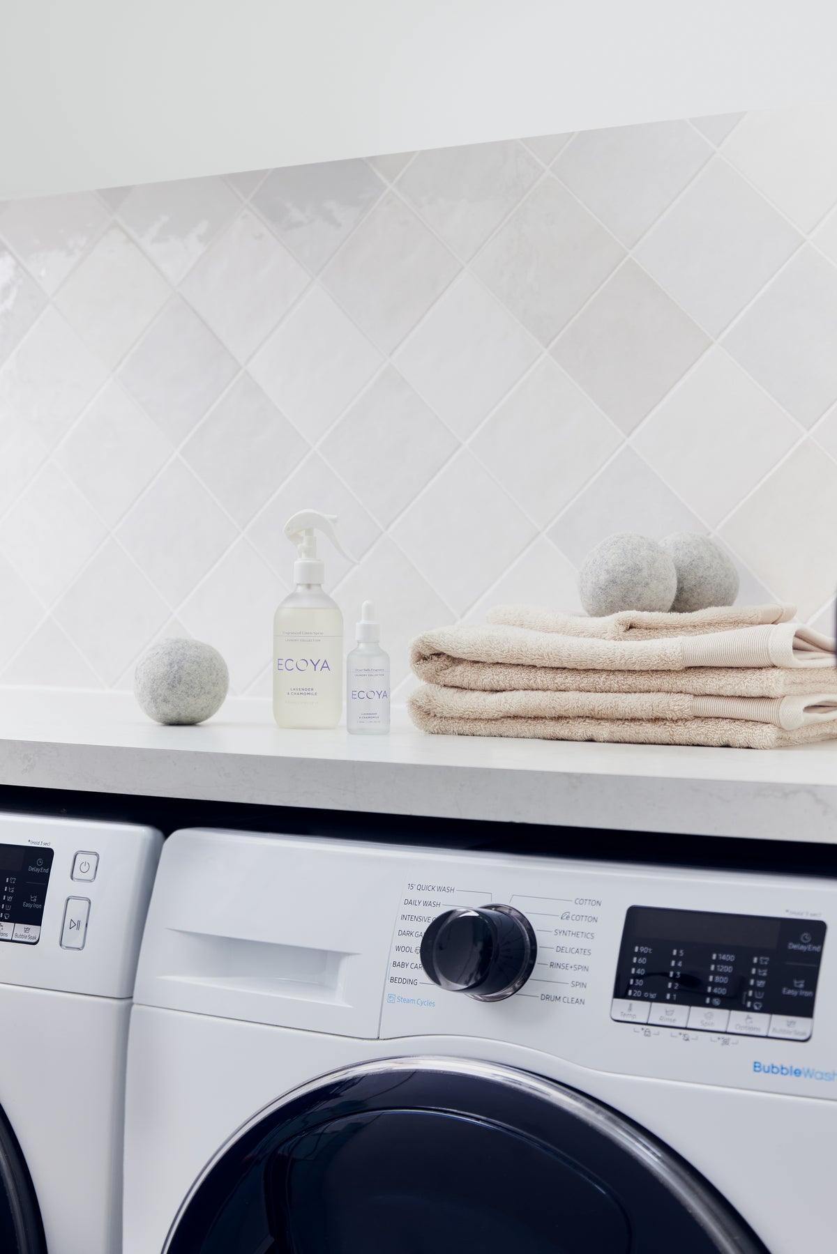 Ecoya laundry liquid, wool dryer balls and folded cream towels on a bench above a washing machine