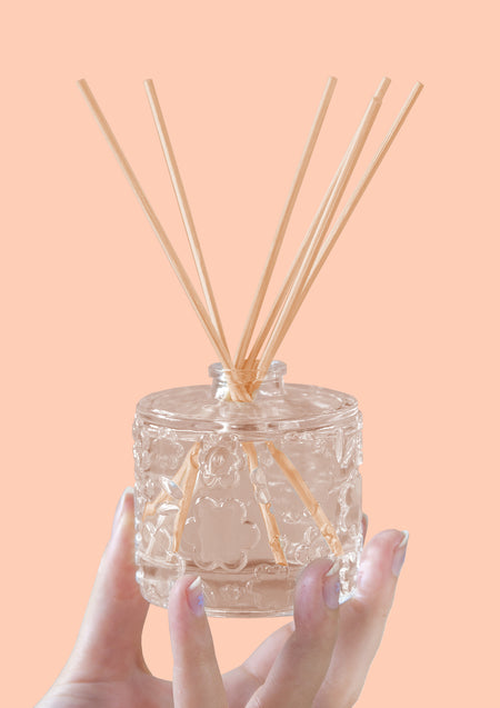 A hand holding the FLWR Fleur D'Oranger reed diffuser against a peach backdrop