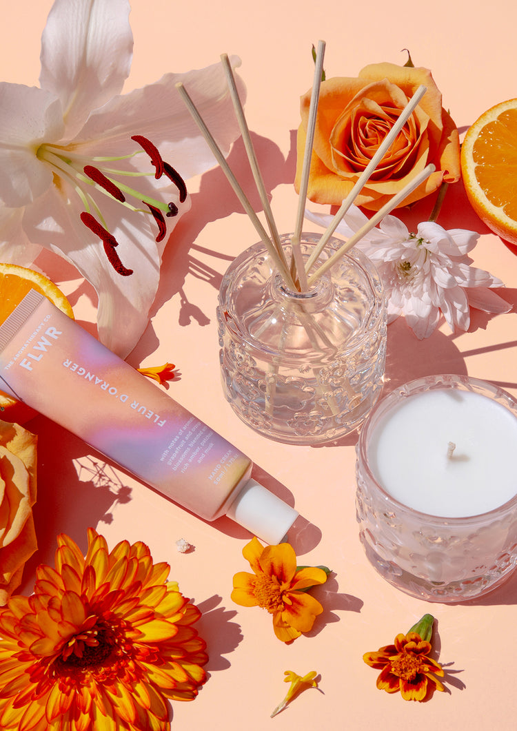 FLWR hand cream tube beside a glass candle and reed diffuser with lilies and orange slices