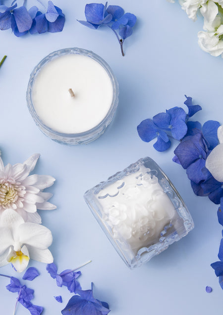 Flat lay of two Forget Me Not candles on pale blue with blue hydrangea petals and white orchids