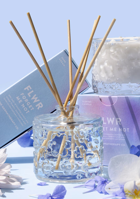 The Forget Me Not diffuser with its blue cartons and a candle behind, blue and white blooms
