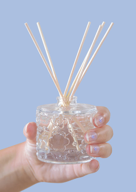 A hand holding the FLWR Forget Me Not reed diffuser against a pale blue backdrop