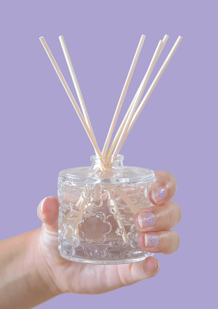 A hand holding the FLWR Purple Reign reed diffuser against a lilac backdrop