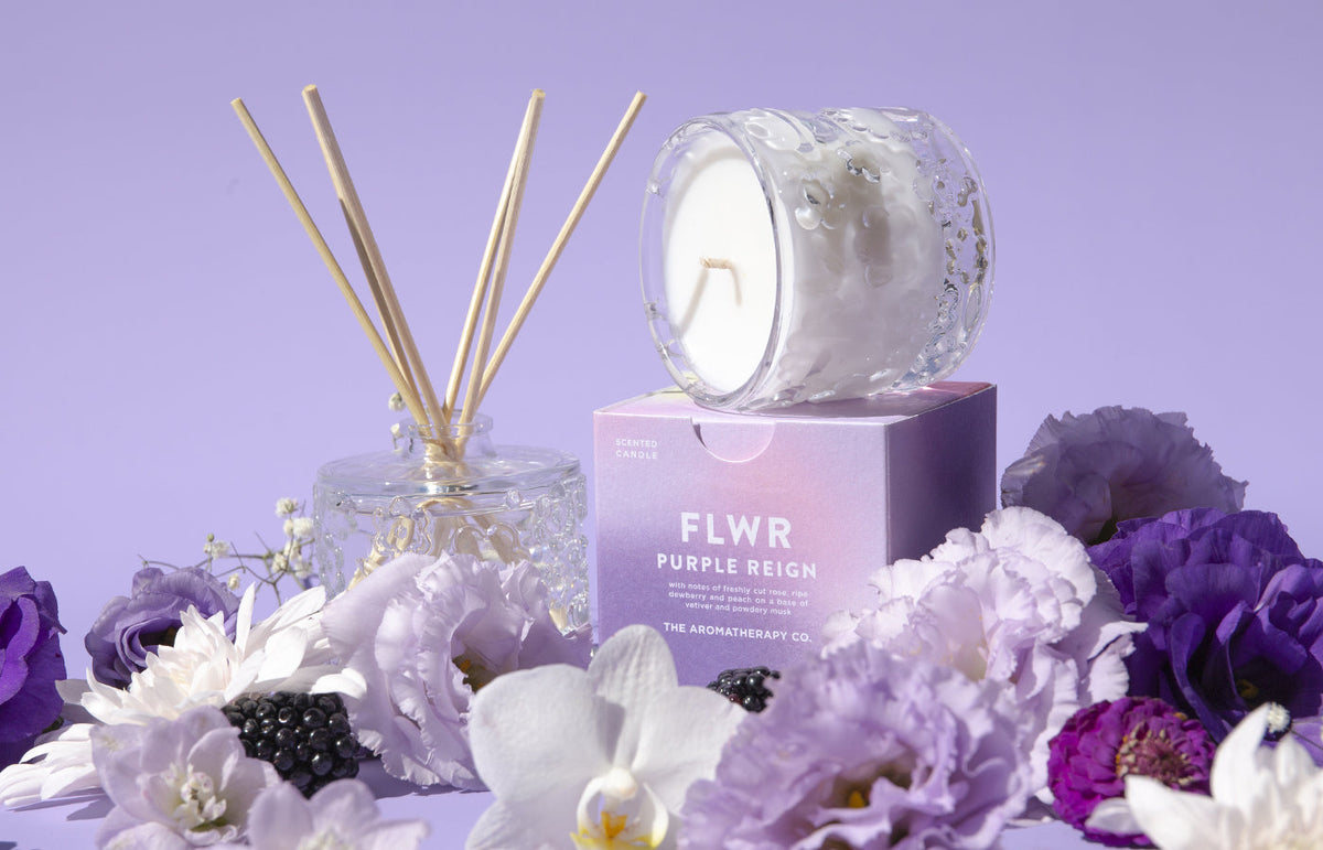 The Purple Reign reed diffuser, carton and candle together among purple blooms and blackberries