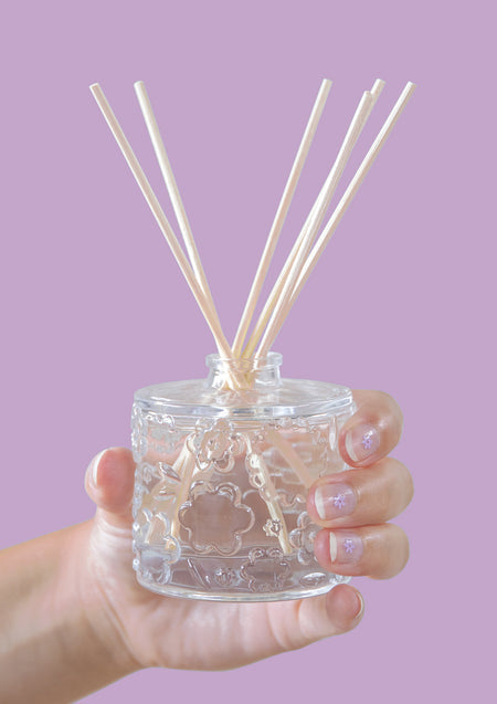 A hand holding the FLWR Sugared Rose reed diffuser against a lilac backdrop