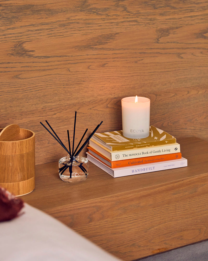 Ecoya reed diffuser on a timber table beside a lit candle and stacked books