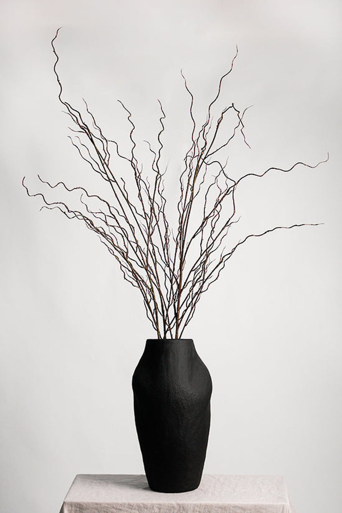 Several twisted willow branches in a matte black vase on a linen-draped plinth