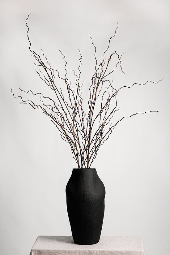 Several twisted willow branches in a matte black vase on a linen-draped plinth