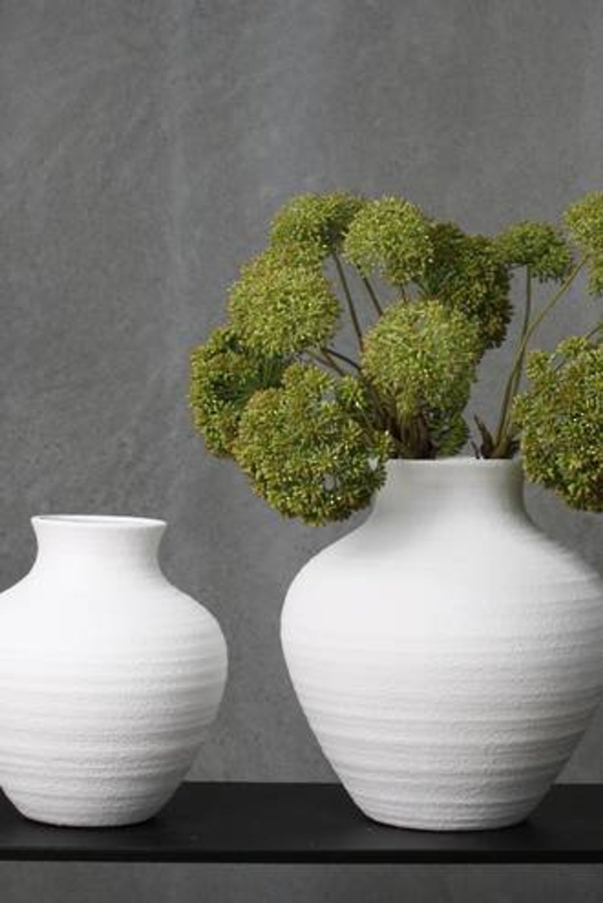 Green sedum sprays in a white ribbed vase beside a smaller vase against grey