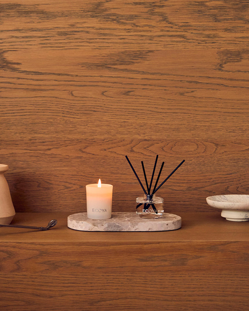 Ecoya reed diffuser and lit candle on a stone tray against a timber wall