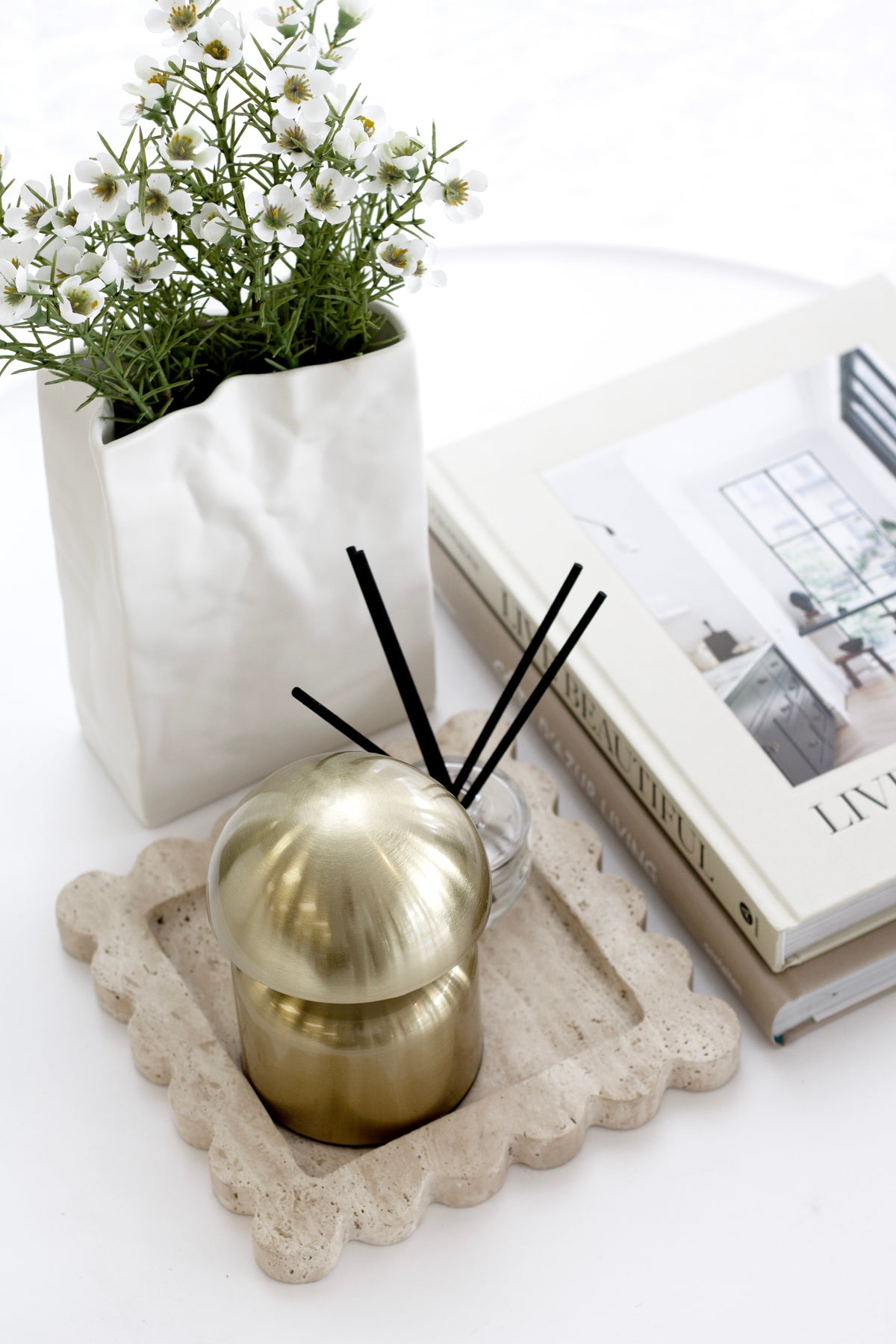 Small white paper bag vase of gypsophila beside a gold diffuser on a scalloped stone tray