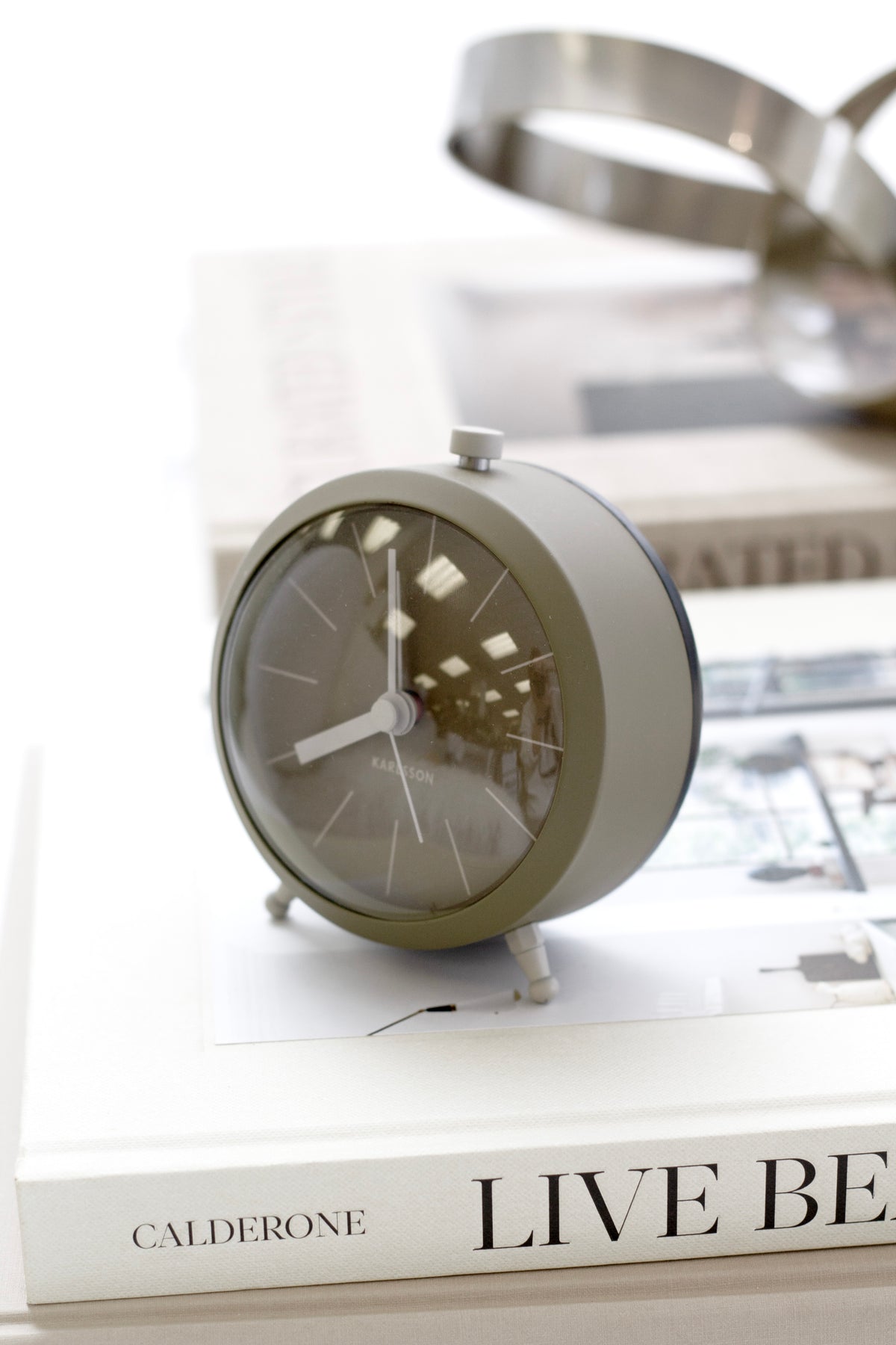 Moss green Karlsson Button alarm clock on a stack of coffee table books beside a silver sculpture