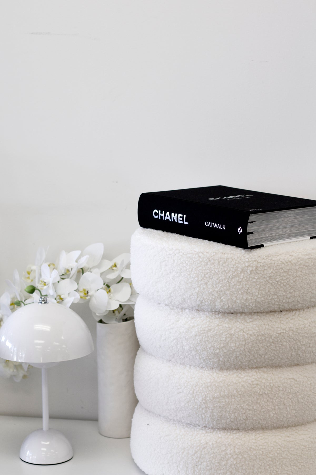White ottoman with a black fashion book on top, beside a dome lamp and white blooms