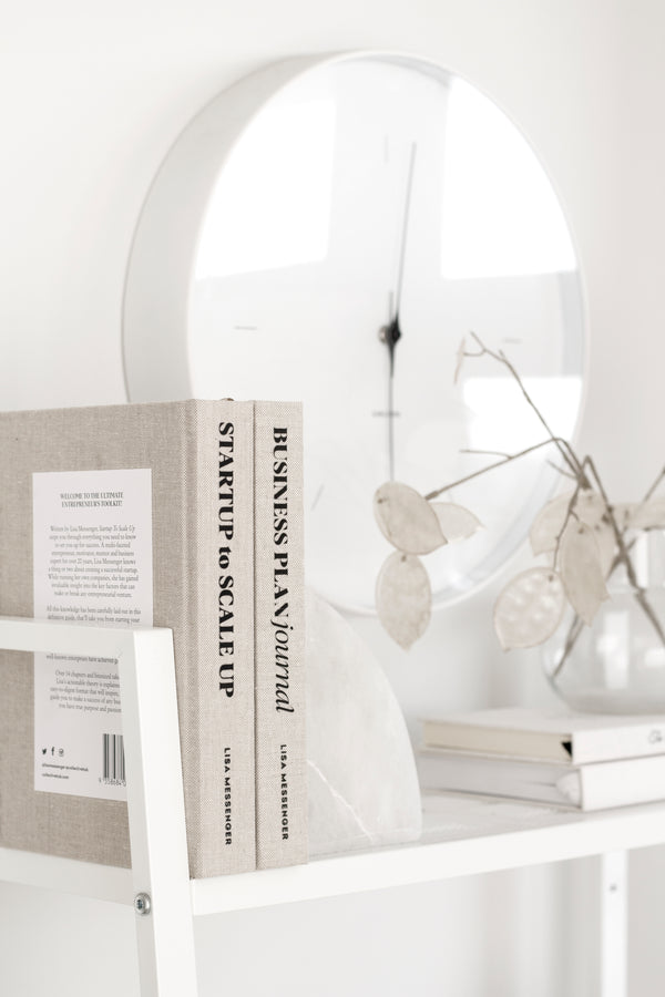 A white shelf with books and a clock on it, perfect for entrepreneurs looking to start up or scale up their business, featuring the Collective Hub Startup to Scale Up Business Plan Journal.