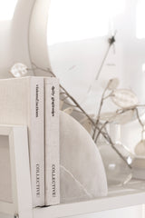 A white shelf adorned with books and a clock, perfect for displaying your favorite literature and keeping track of time as you plan your goals and document them in your Visions & Actions Journal by Collective Hub.
