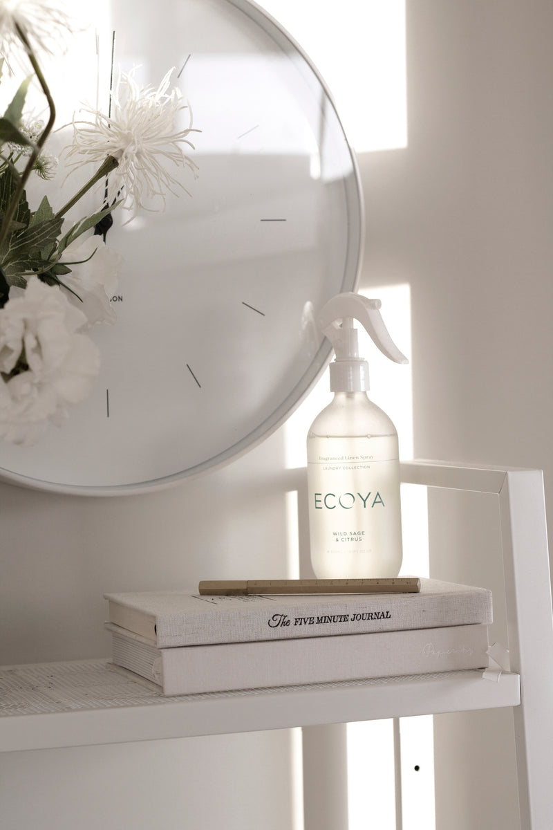 A bottle of Ecoya Laundry Linen Spray is sitting on a shelf next to a clock.