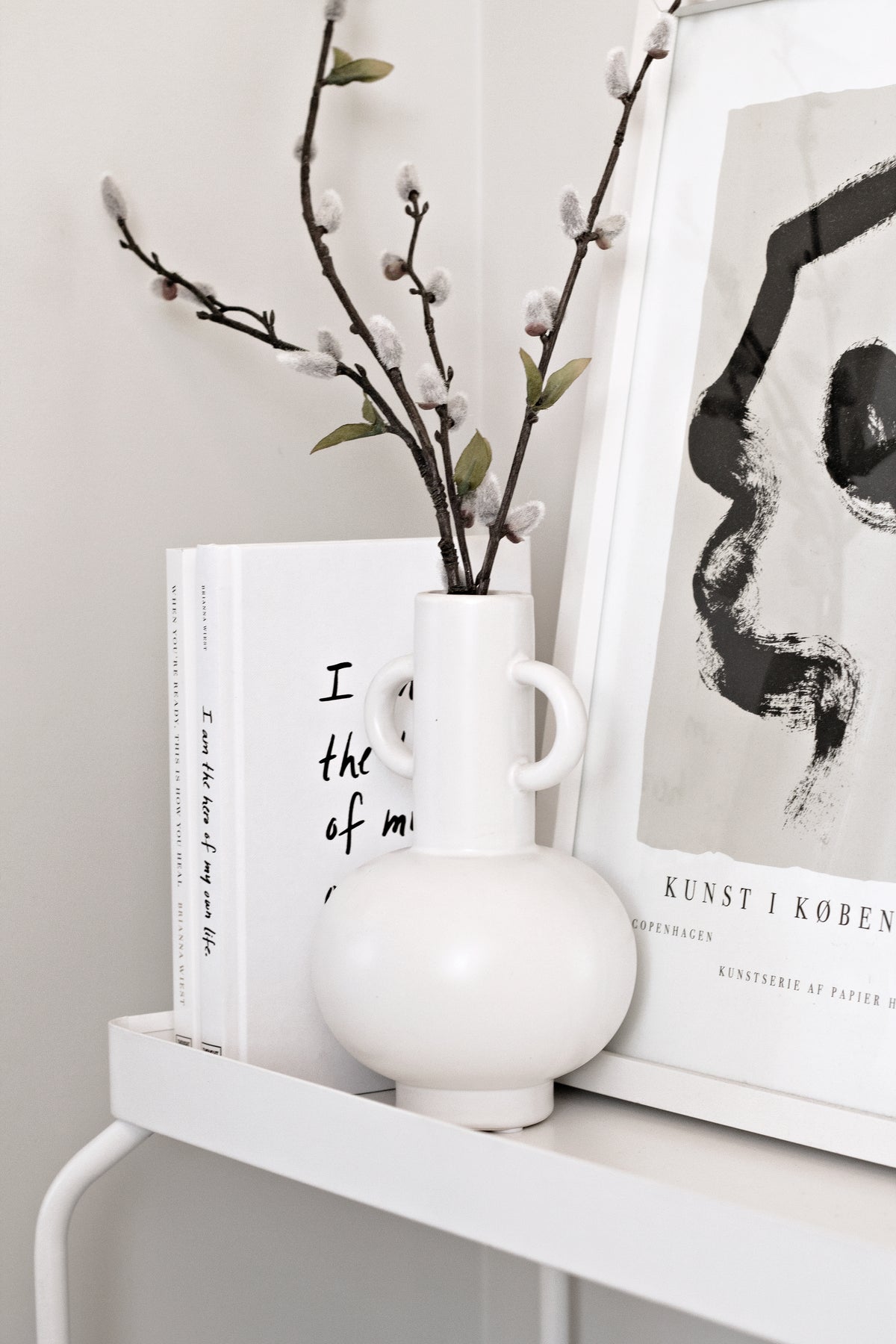 A minimal stack of Brianna Wiest’s books, including “I Am the Hero of My Own Life,” styled between travertine bookends on a clean white shelf.