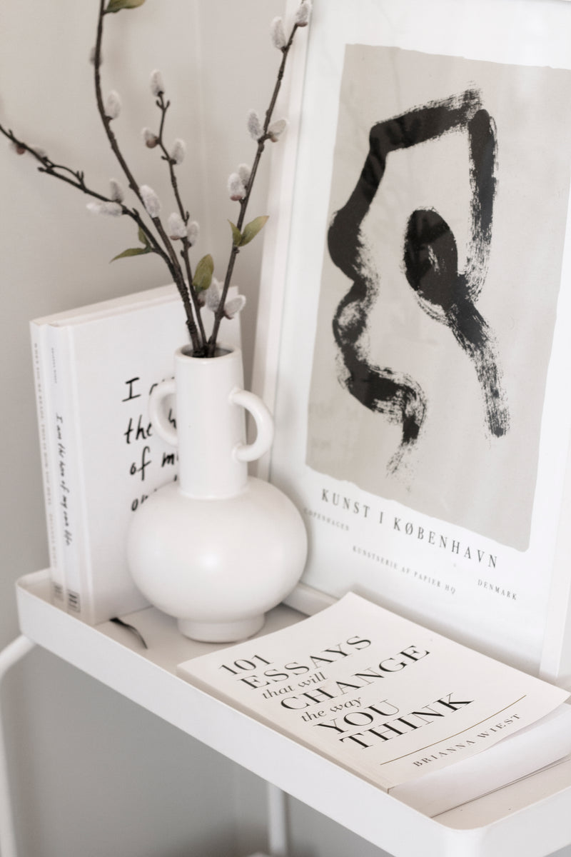 A white shelf showcasing books, including "101 Essays That Will Change The Way You Think" by Brianna Wiest, accompanied by a vase and an art print from Thought Catalog.