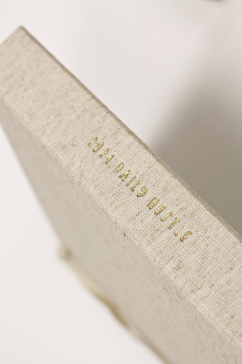 A 2024 DAILY HUSTLE PLANNER. OATMEAL with gold lettering on a white surface by Write To Me.
