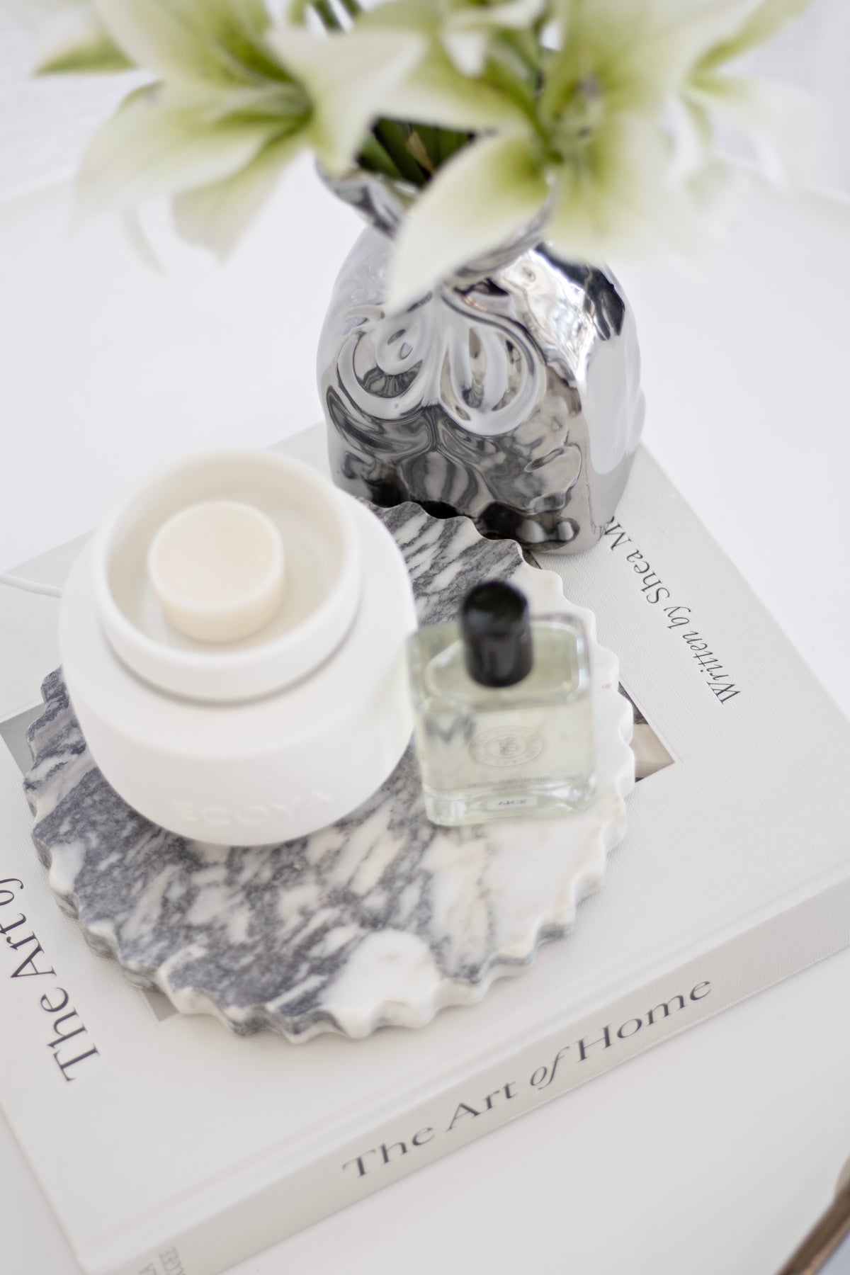 Cream lilies in a marbled vase with a candle, fragrance bottle and books