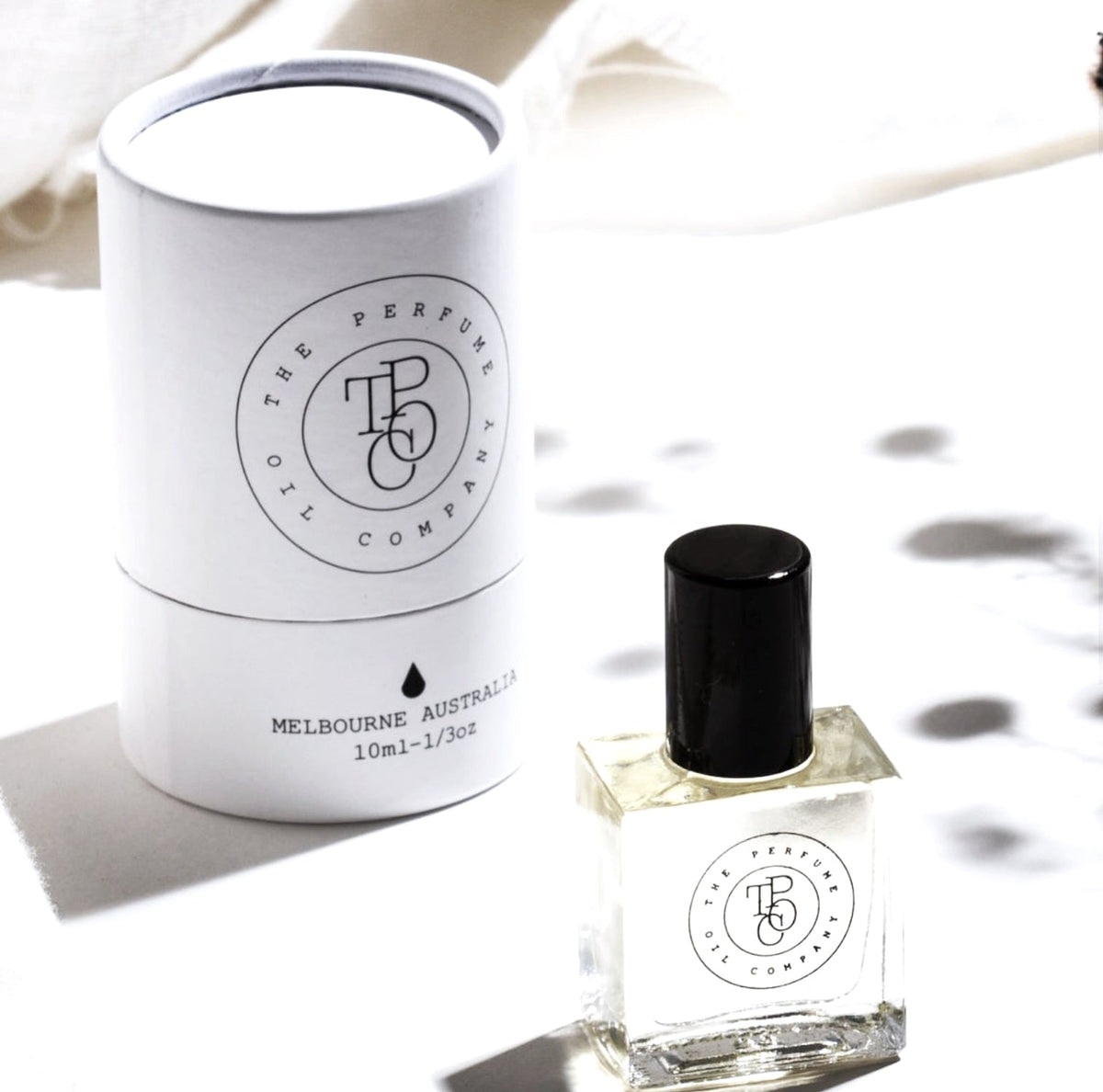 White perfume oil tube carton behind a roll-on bottle with soft leaf shadows