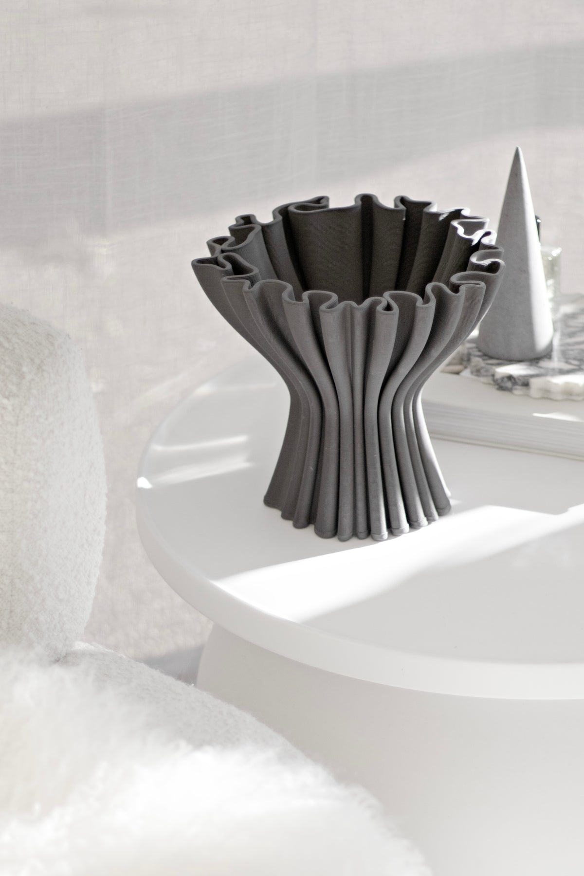 Cleo compote vase in dark grey on a round white table beside a marble cone ornament, with a boucle chair alongside