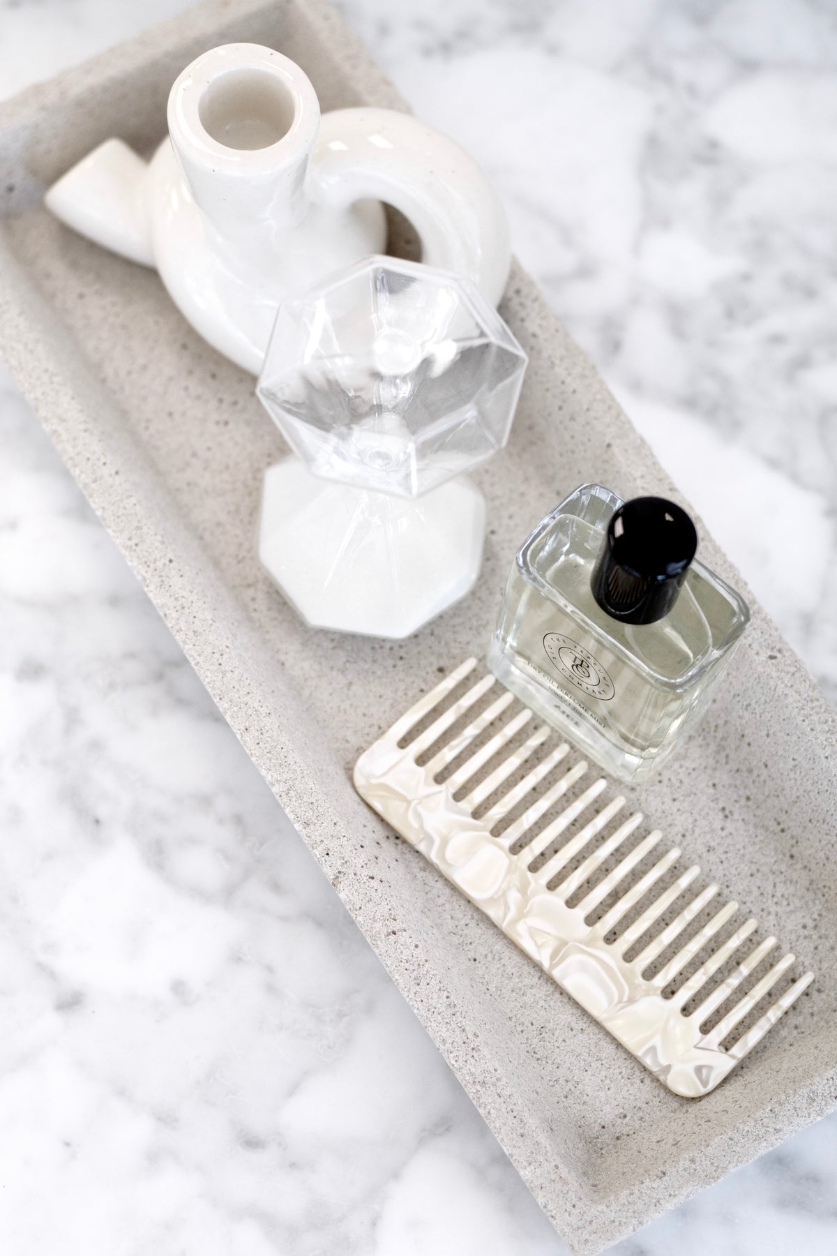 Overhead of a speckled concrete tray holding the knot holder, an hourglass, a perfume bottle and a comb