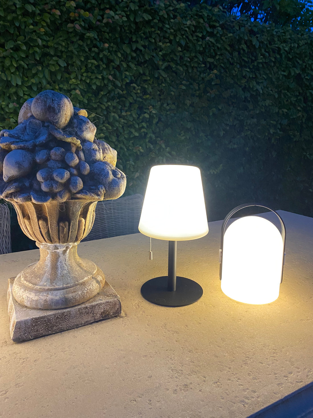 Hampton LED lantern lit on an outdoor table at dusk beside a stone urn planter