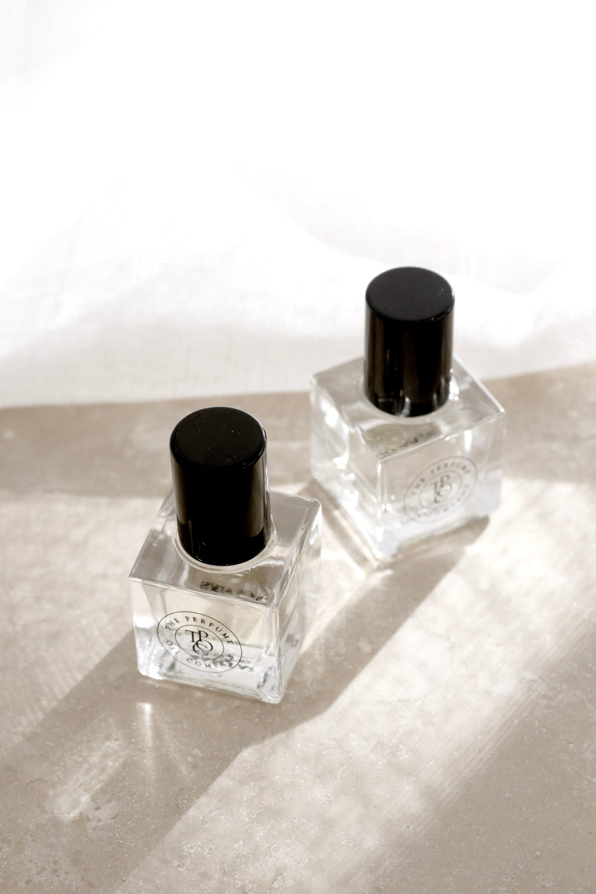 Two clear cube roll-on perfume oil bottles with black caps on a pale surface in soft sunlight