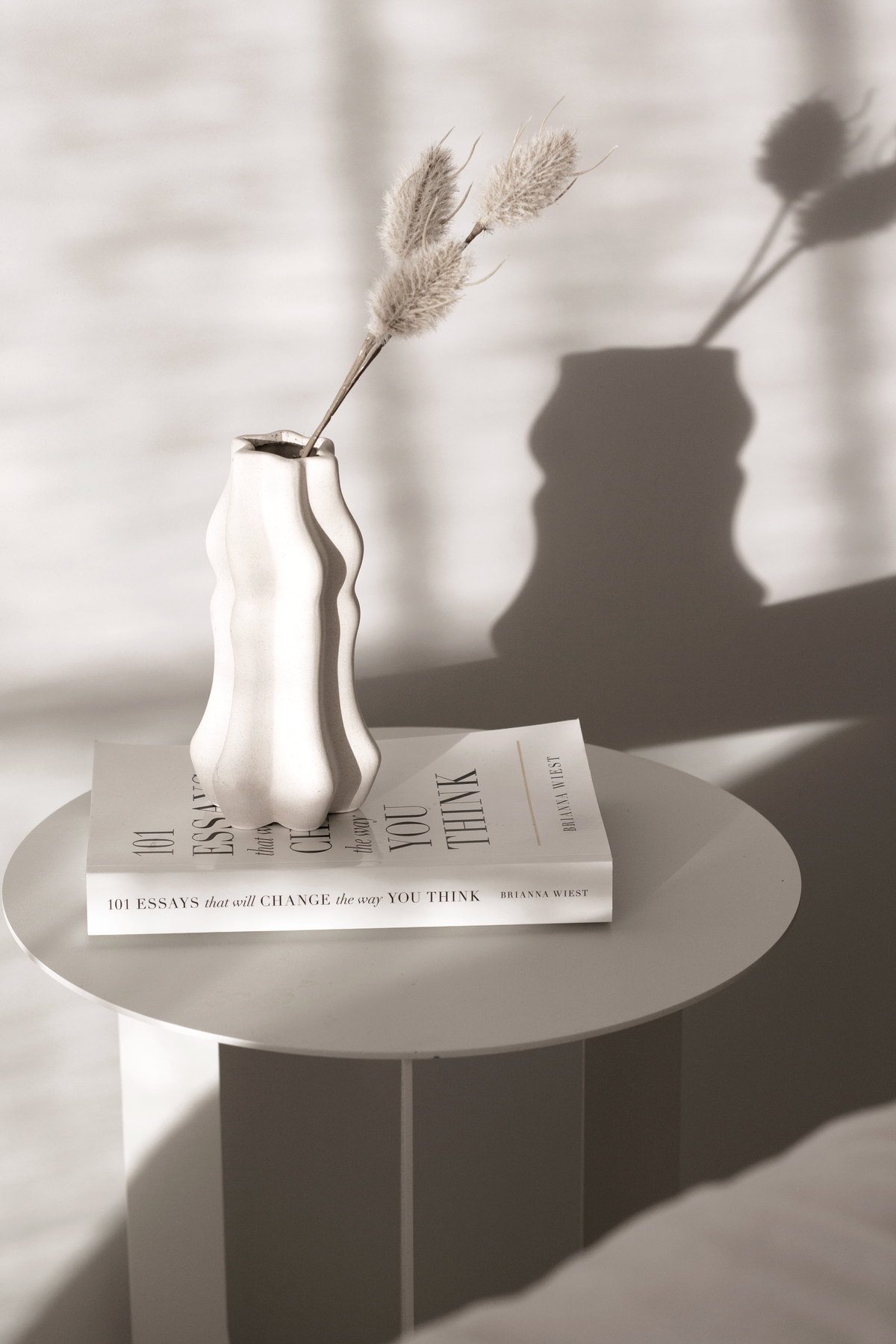 The 101 Essays paperback on a ribbed side table beside a bed, with a wavy vase of bunny tail grass