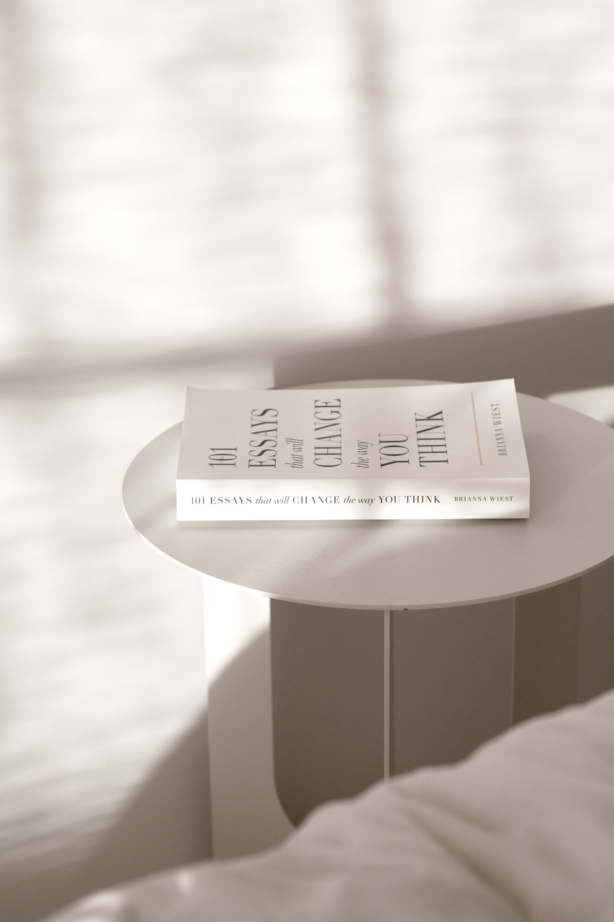 The 101 Essays paperback lying on a ribbed side table beside a bed, sheer curtain behind