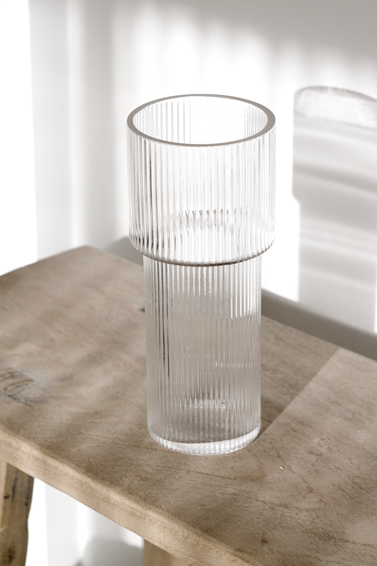 Empty ribbed glass vase on a timber console beside a sheer curtain