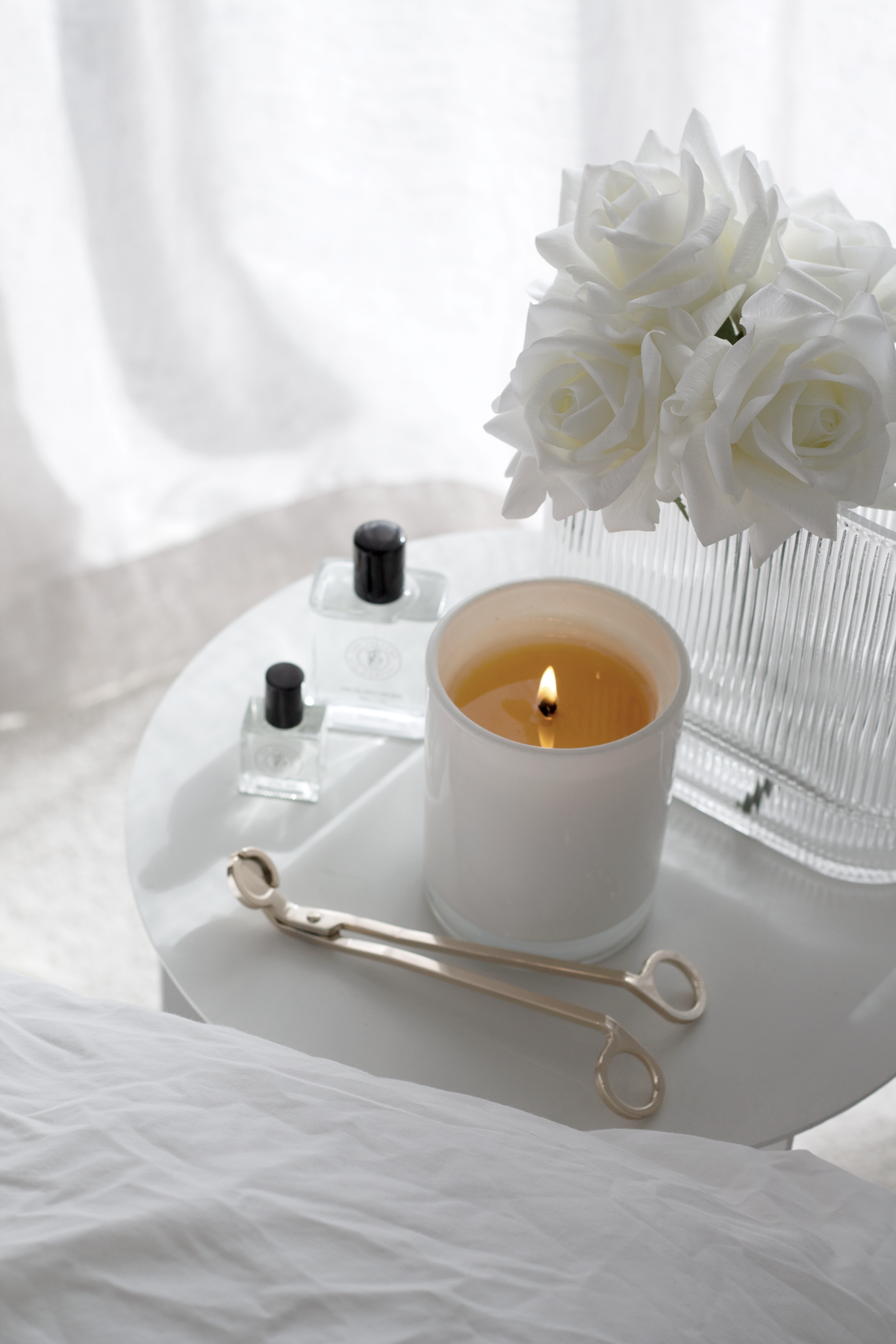 Gold wick trimmer on a stone tray with a lit candle and white roses on rumpled linen