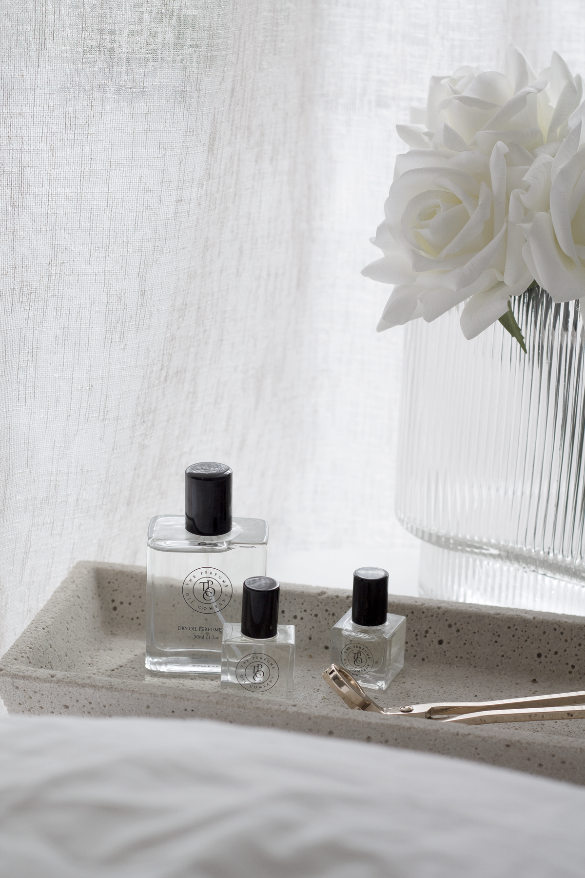 Gold wick trimmer on a stone tray with fragrance bottles and white roses in a ribbed vase