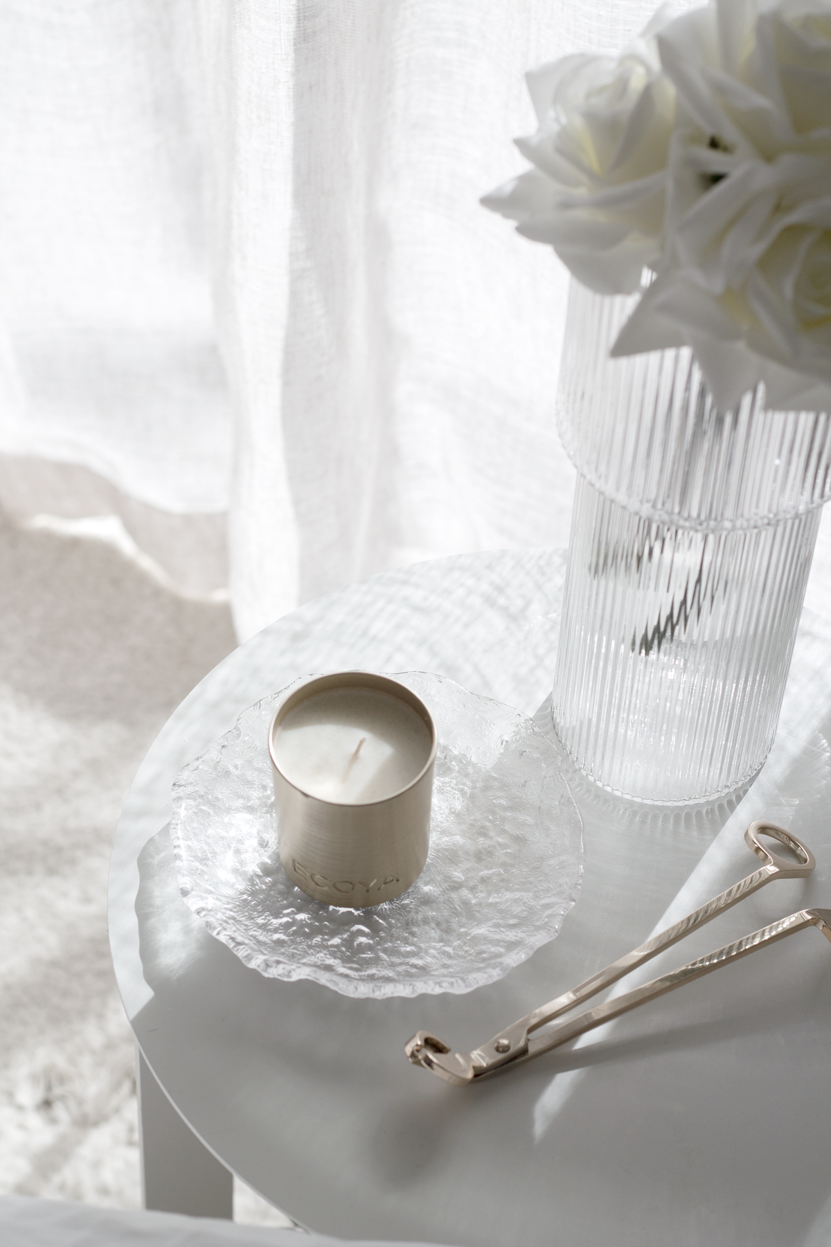 Gold wick trimmer on a round tray beside a candle and white roses in a ribbed vase