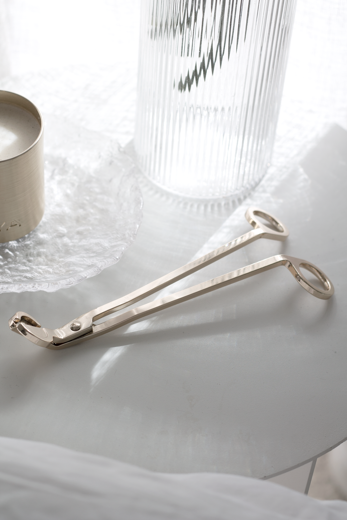 Silver wick trimmer on linen beside a candle and a ribbed glass vase in soft light