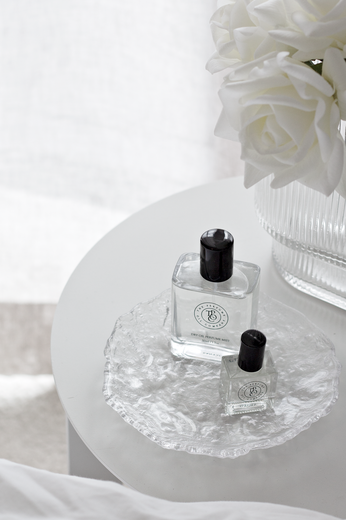 Glacier glass tray holding a perfume bottle on a round table, white roses behind