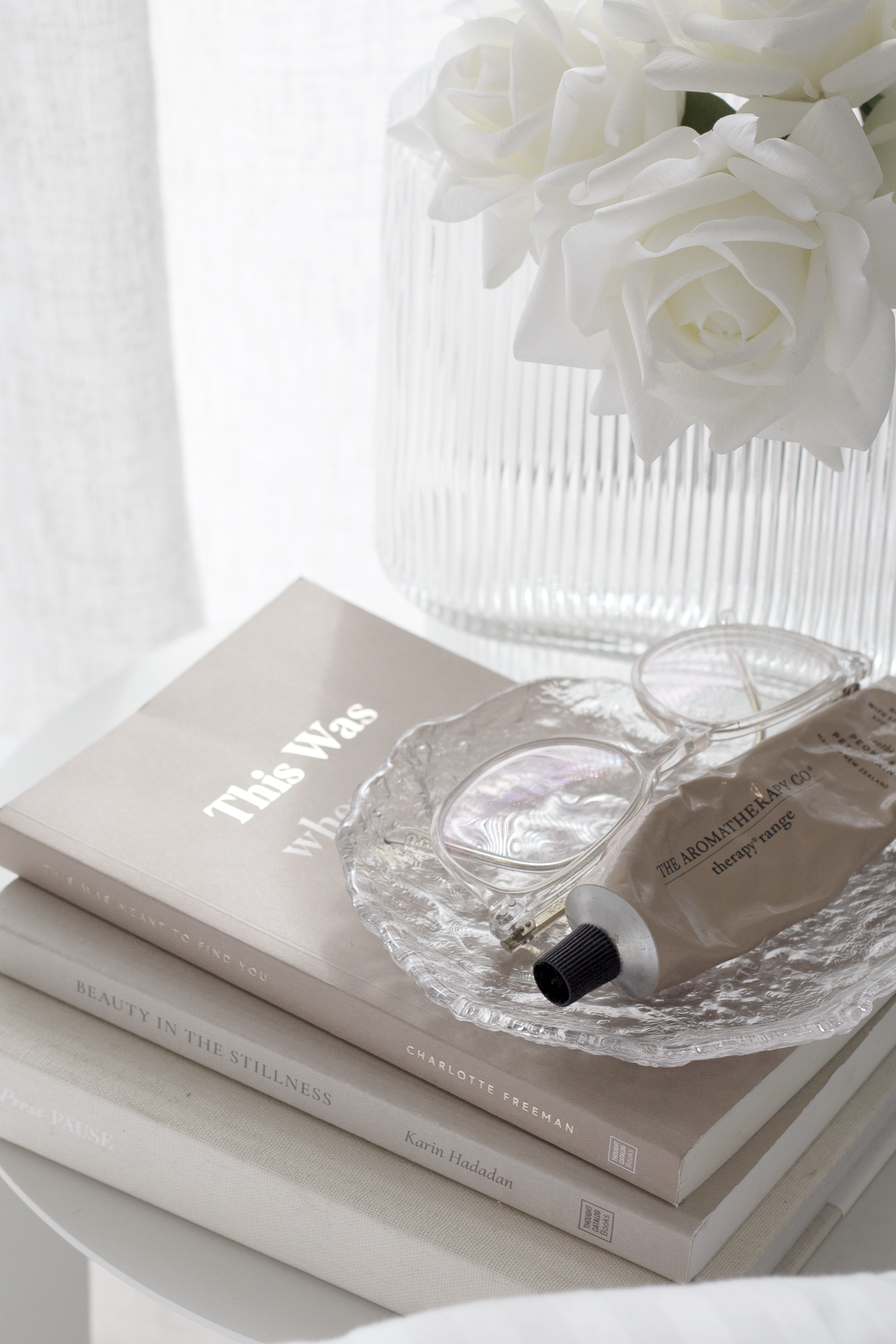 Hand cream tube resting in a glass dish on stacked books beside white roses