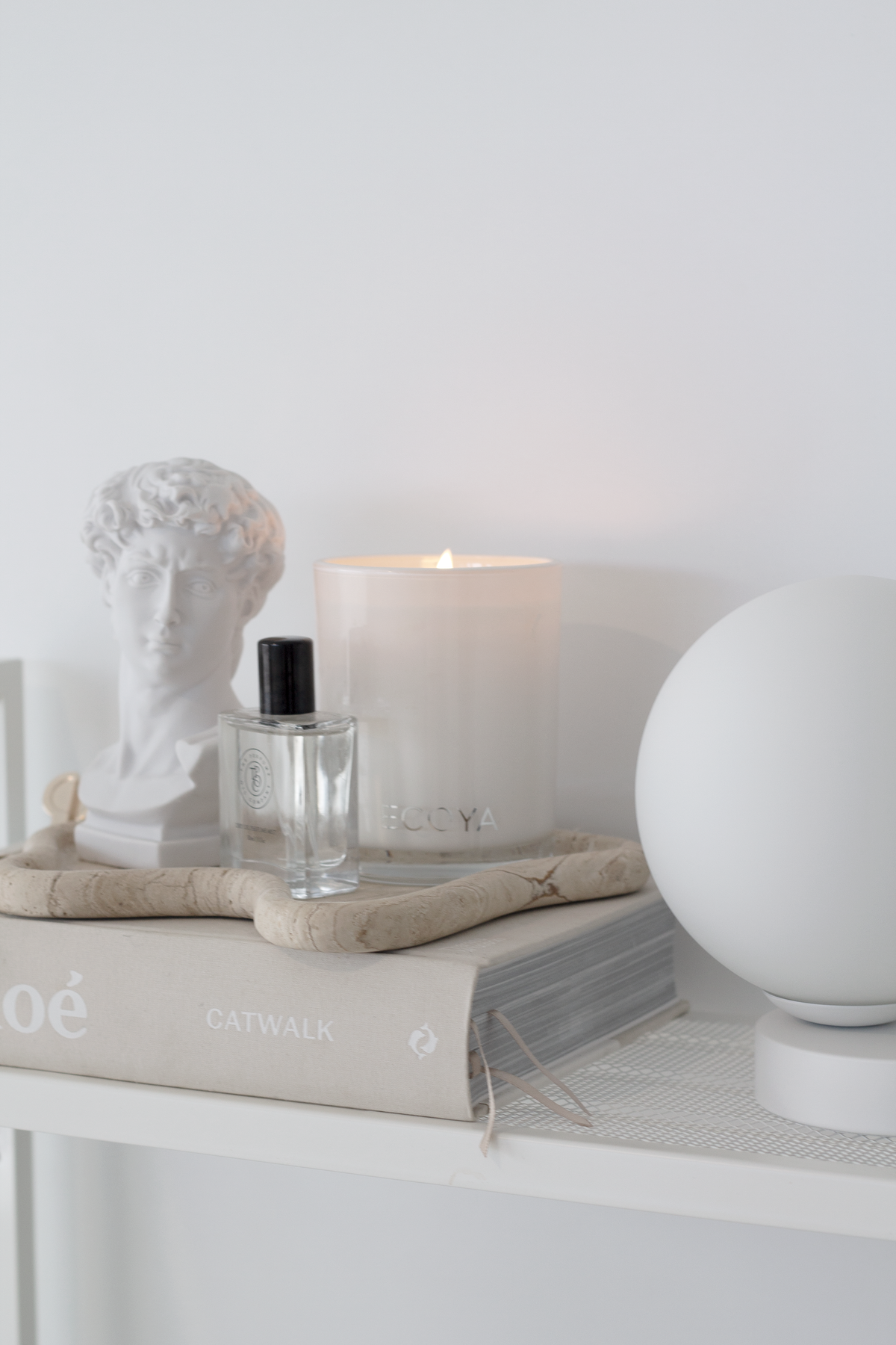 Yuri Table Lamp on a white shelf beside a candle, a perfume bottle and a bust sculpture