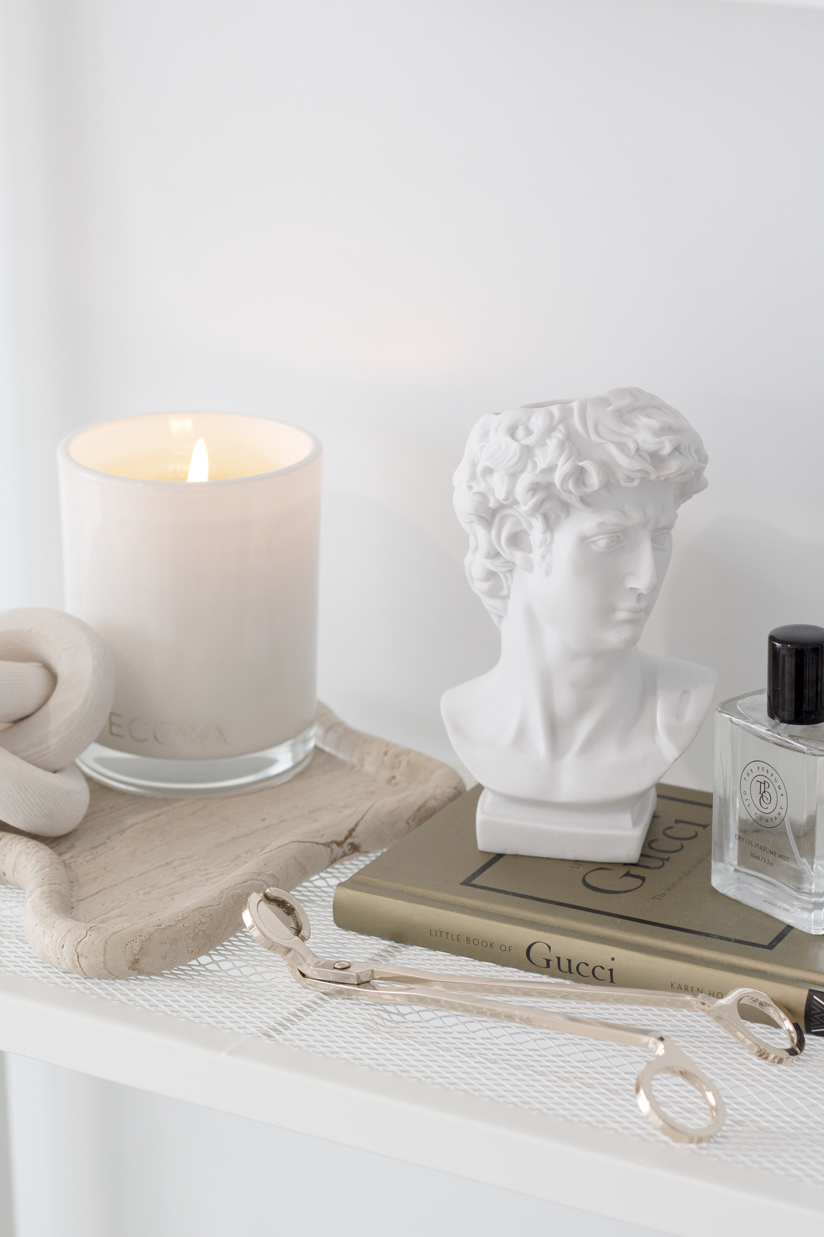 Gold wick trimmer on a shelf with a lit candle on stone, a bust sculpture and a fragrance bottle