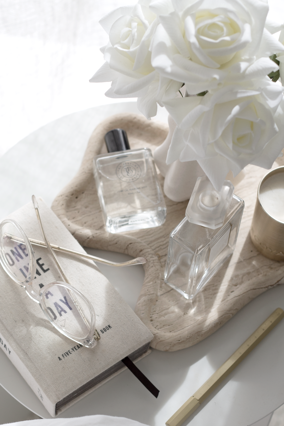 Soft morning flatlay featuring a five-year journal with reading glasses, two clear perfume bottles, a bunch of white roses in a sculptural vase, a lit candle, and a gold pen, all styled on a textured tray in natural light