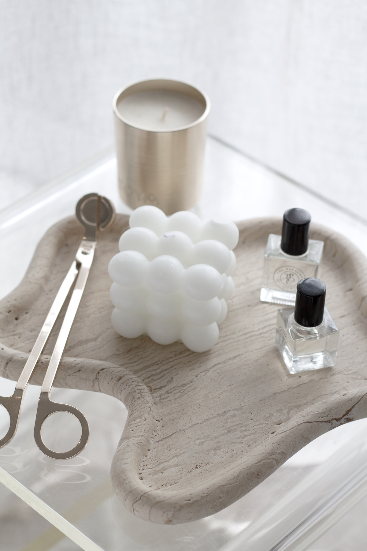 Gold wick trimmer on a travertine tray with a bubble candle and fragrance bottles