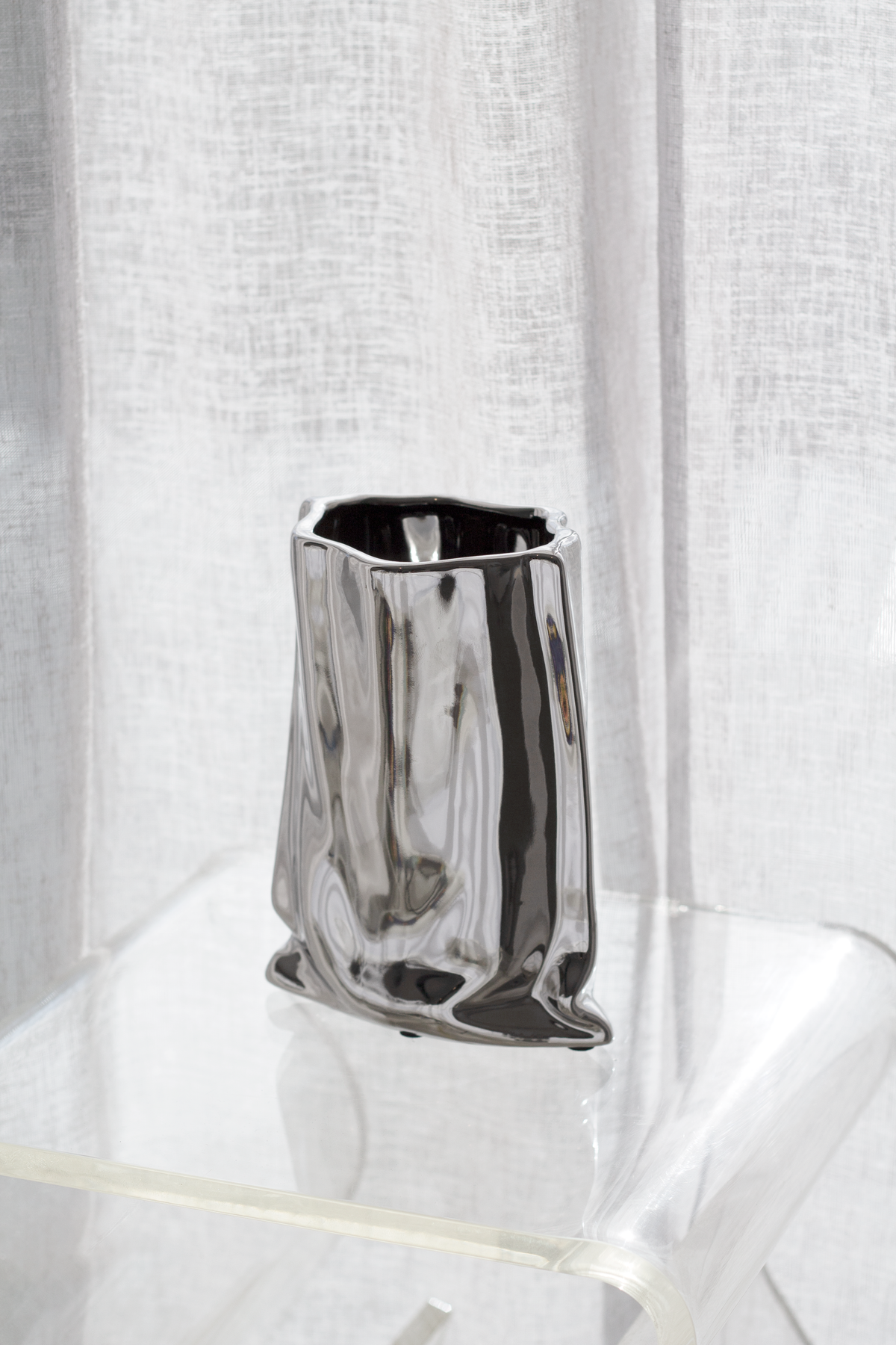Silver metallic bag vase on a clear acrylic table by a sheer curtain