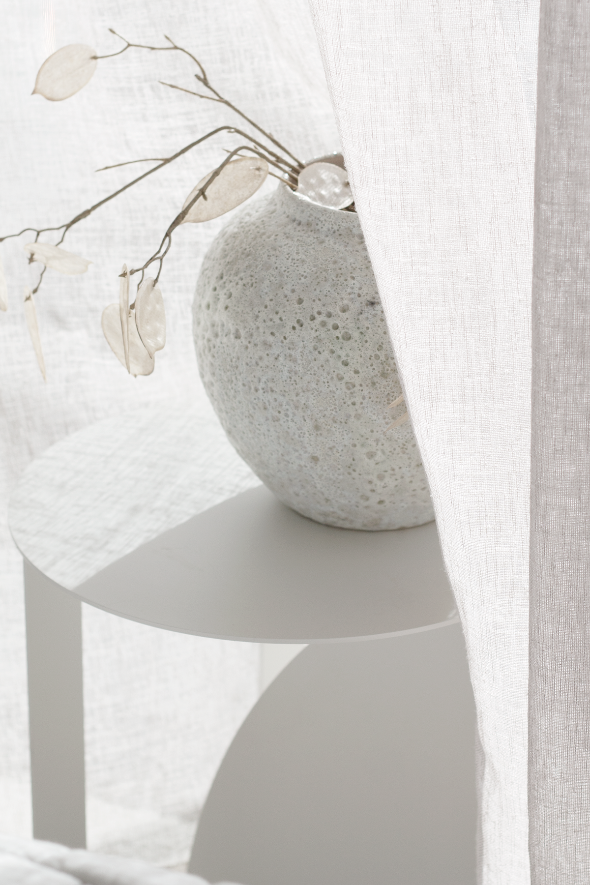 Short Seafoam vase of honesty stems on a round side table beside a sheer curtain